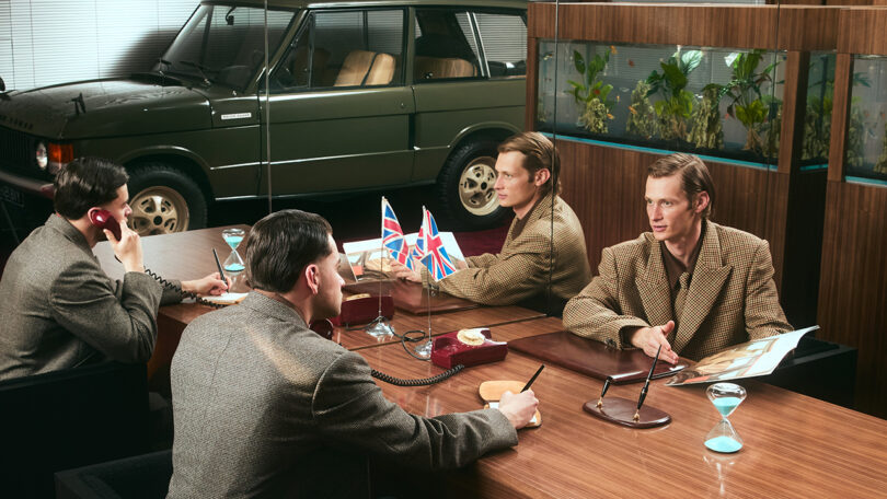 Four men in brown checkered jackets sit at a wooden table with British flags, phones, hourglasses, and documents; a green car and fish tanks are visible in the background.