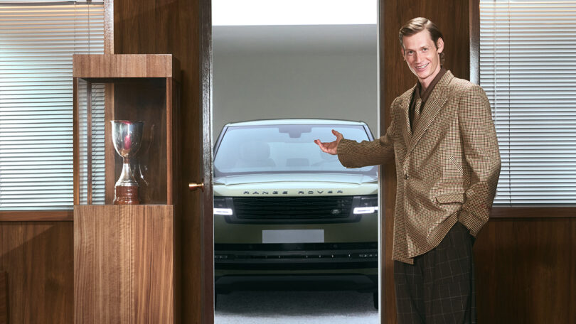 A man in a checked blazer stands in an office, gesturing toward a green Range Rover parked outside an open door. A trophy is displayed in a glass case nearby.