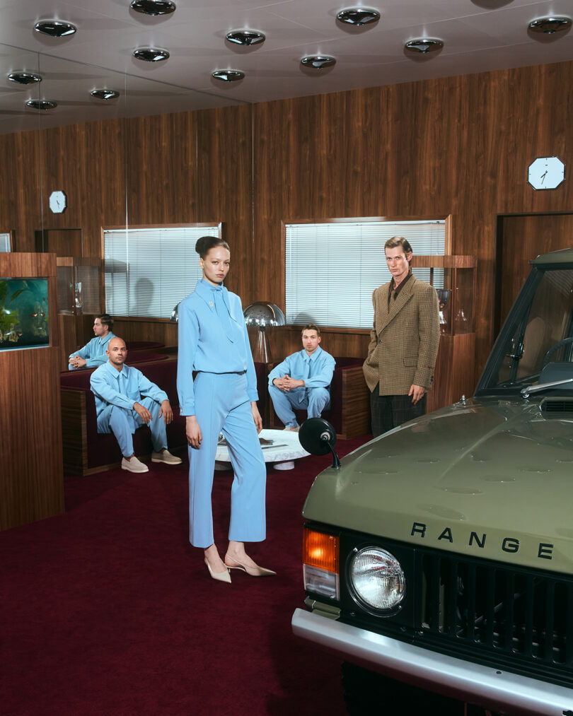 Four people in an office with wood-paneled walls and red carpet; three wear light blue outfits, one wears brown. A green Range Rover is parked indoors in the foreground.