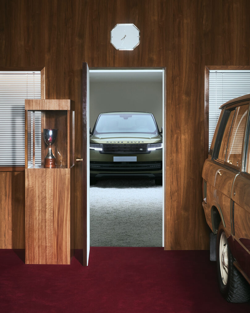 A modern SUV is visible through a doorway in a wood-paneled room with red carpet, a trophy case, and part of a classic car.