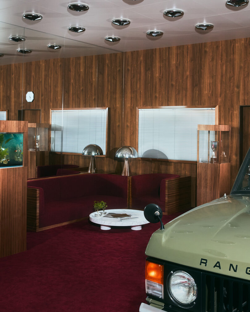 A vintage-styled room with wood paneling, red carpet, modern furniture, dome lamps, and part of a green Range Rover visible in the foreground.