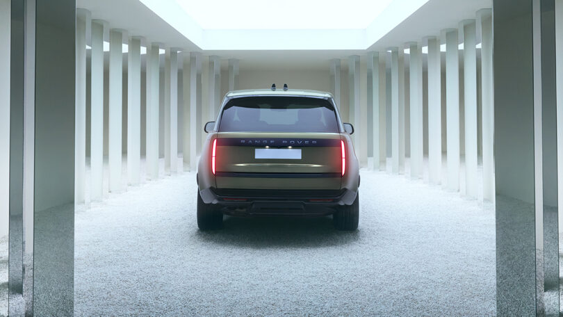 A rear view of a Range Rover SUV parked on a gravel floor between rows of white columns in a modern, minimalist setting.