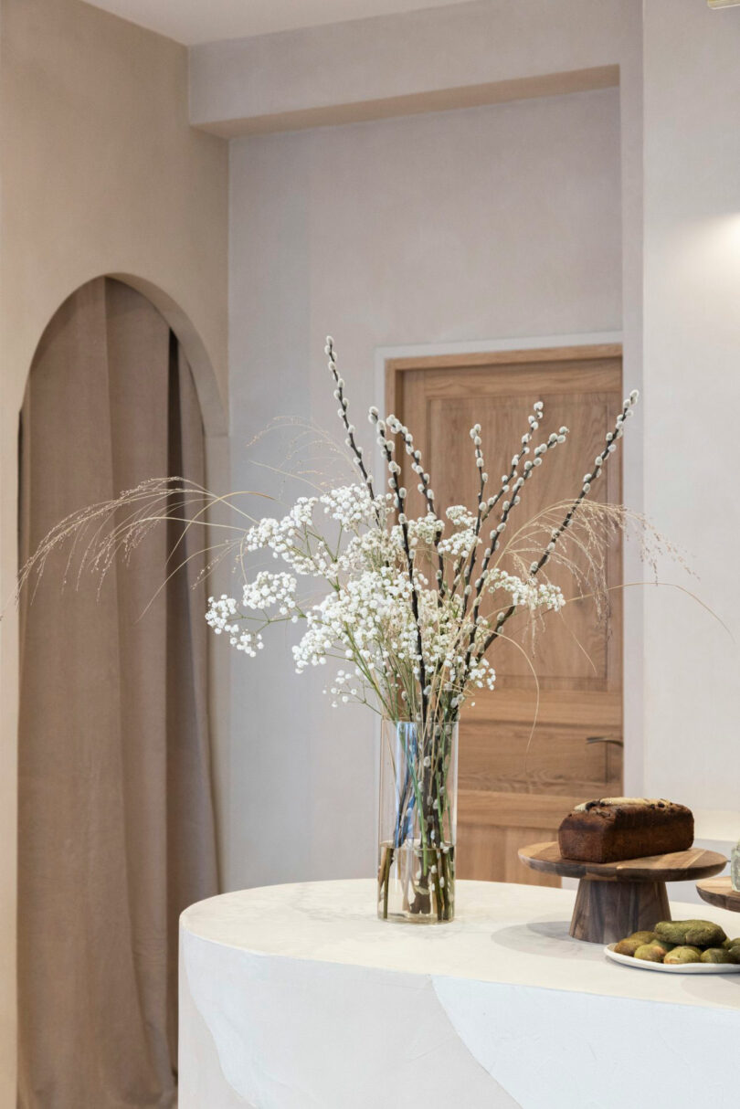 A glass vase with white flowers and tall stems sits on a round, light-colored table beside a cake and a plate of cookies in a minimalist room with neutral walls and a wooden door.