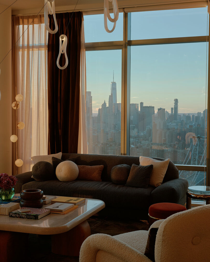 Cozy living room with a dark sofa, various cushions, a table with books, and pendant lights. Large window reveals a city skyline at sunset.
