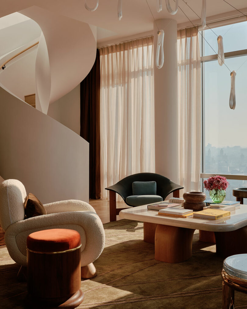Modern living room with plush chairs, a large square coffee table, and a spiral staircase. Sunlight filters through sheer curtains. Pink flowers and books adorn the table.