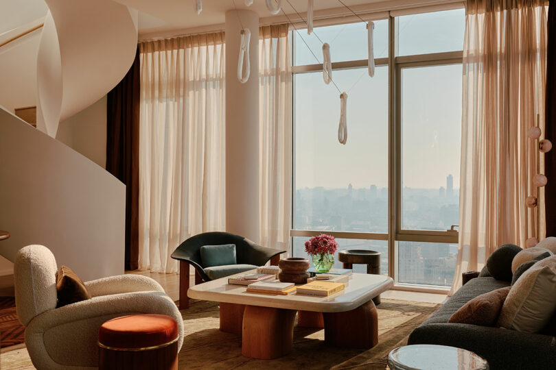 Modern living room with a large window, spiral staircase, and contemporary furniture, including a white table and curved chairs. Beige curtains partially drawn, and books and flowers on the table.