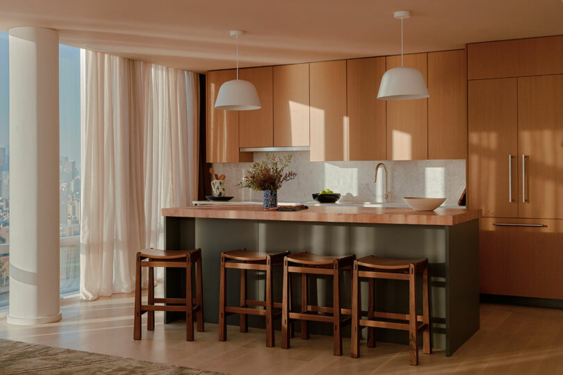 Modern kitchen with light wood cabinets, an island with a wooden countertop, and four stools. Large windows with sheer curtains provide natural light. White pendant lights hang above the island.