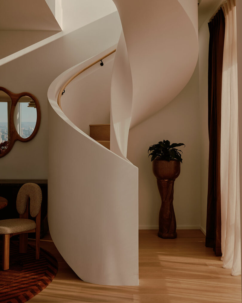 A modern spiral staircase with white walls and a wooden handrail. Nearby, there's a round table, chairs, a mirror on the wall, and a potted plant in a wooden stand. Natural light fills the room.