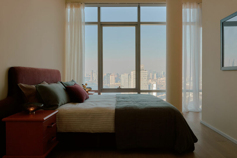 A bedroom with a large bed, pillows, and a side table. A tall window fills the room with natural light, offering a view of a city skyline.
