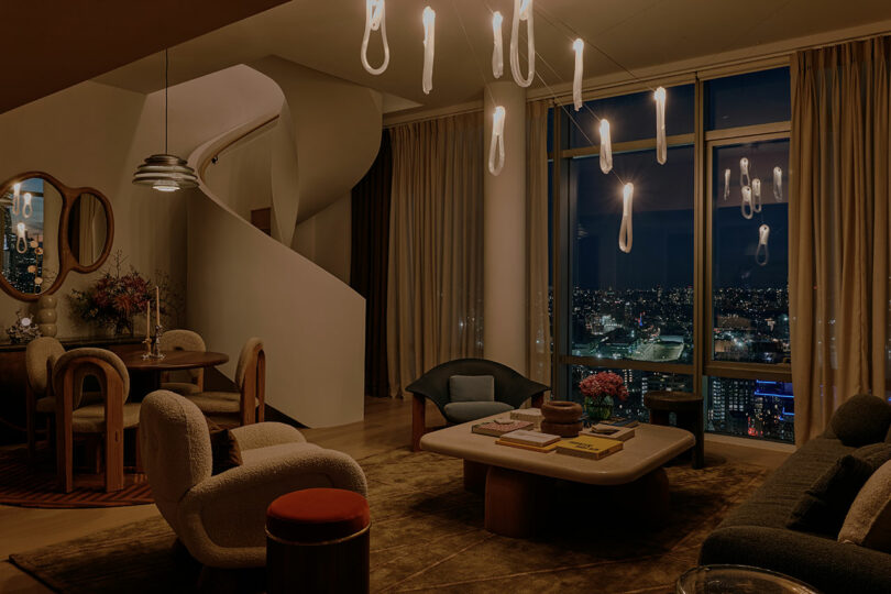 Modern living room with night city view. Features a spiral staircase, eclectic seating, a coffee table with books, pendant lights, dining set, and large windows with curtains.