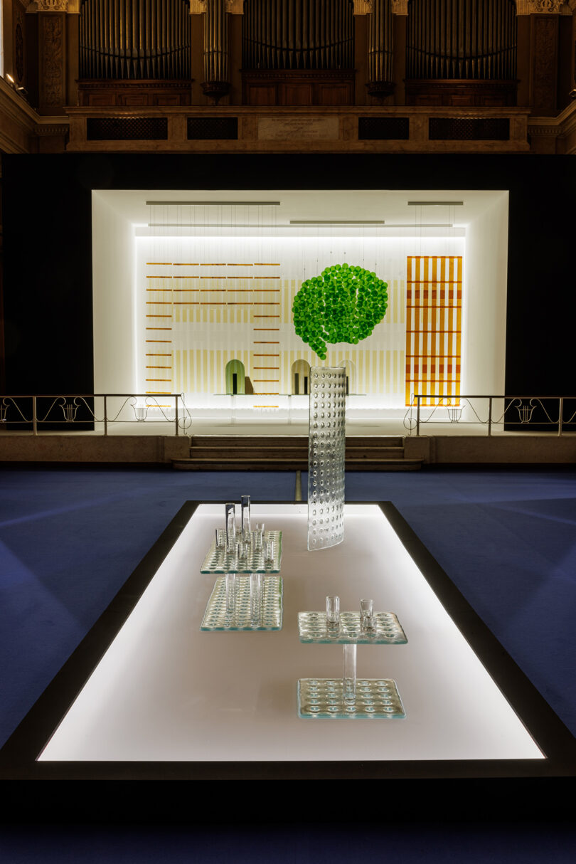 A modern art installation featuring glass objects on a lit platform in the foreground, with a geometric wall display and green circular plant in the background.