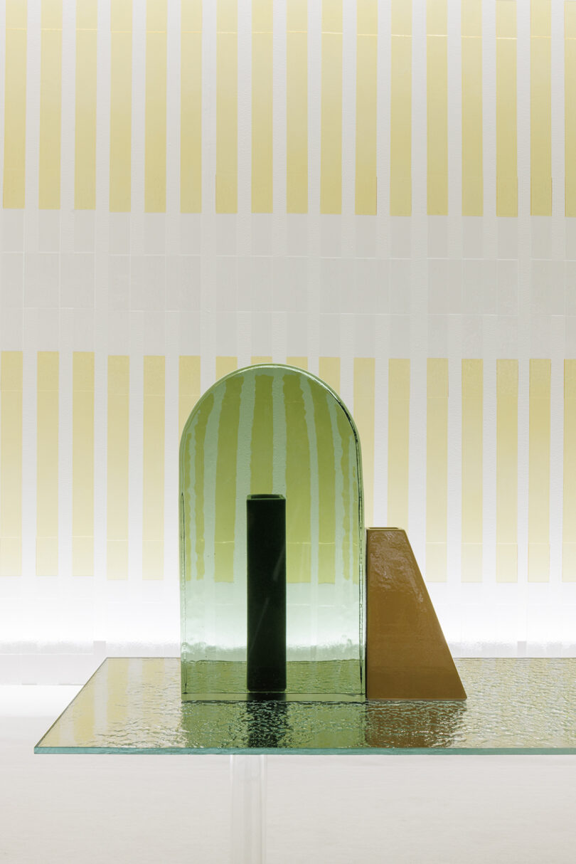 A geometric glass and metal sculpture with a green arch, black cylinder, and yellow wedge sits on a textured glass surface against a striped yellow and white background.