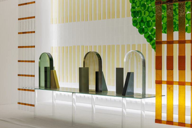 A modern glass table displays geometric sculptures with arched and rectangular shapes, set against striped and circular patterned translucent panels.