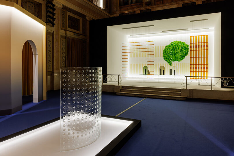 A modern art installation in a grand hall features a transparent cylindrical sculpture with circular cutouts and a stage backdrop with geometric patterns and a green tree design.