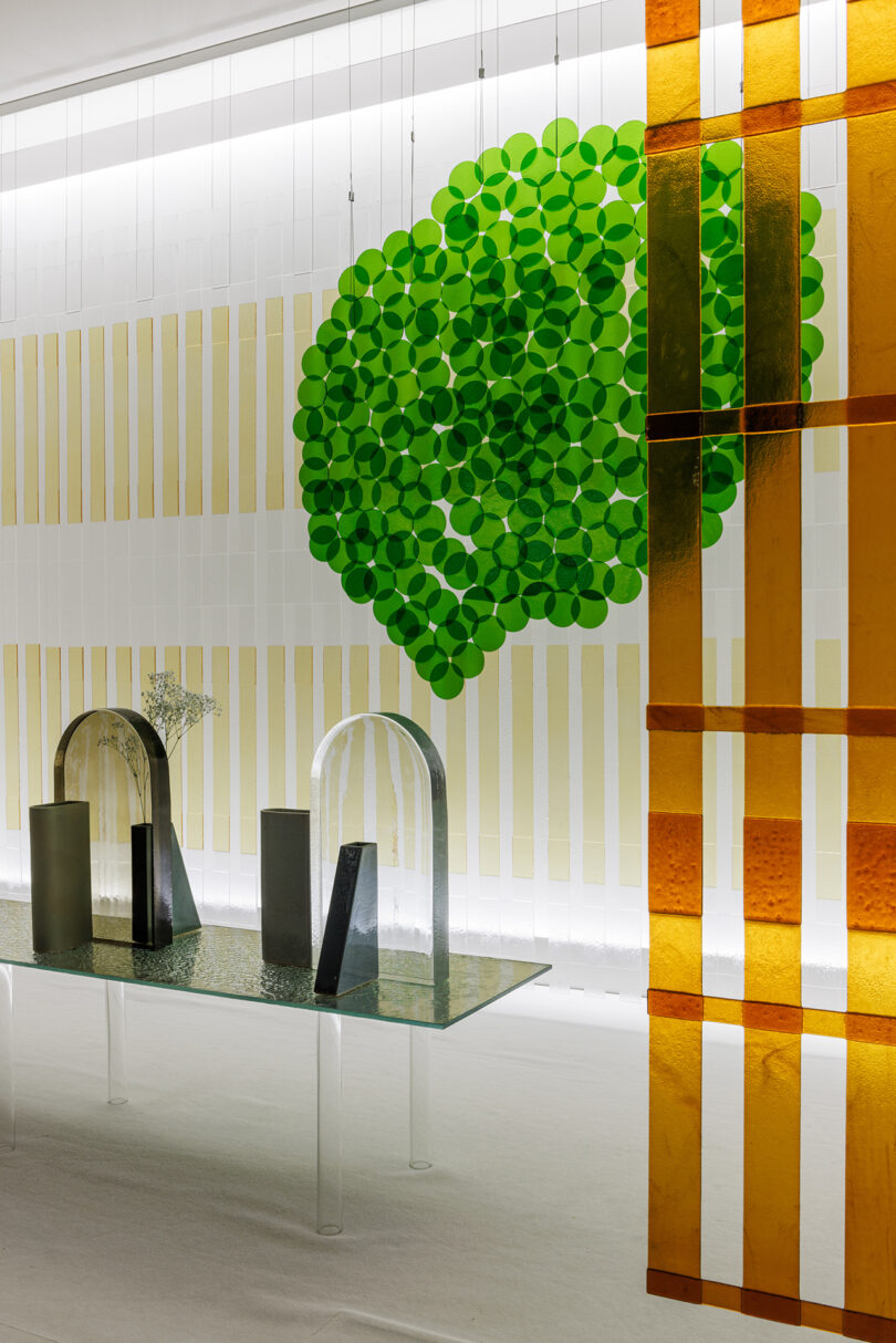 A modern interior with a glass table holding geometric sculptures, a green circular wall art piece, and amber vertical panels in the foreground.