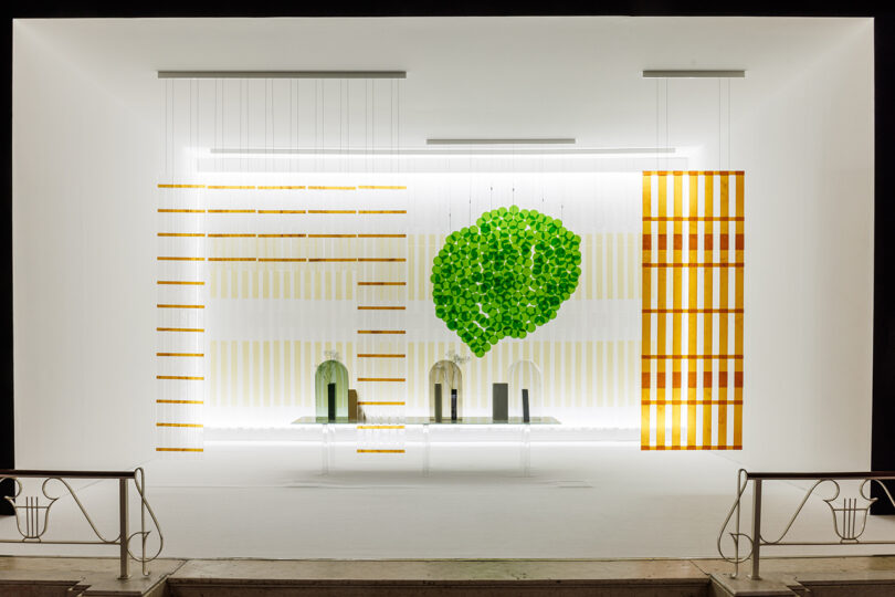 A modern window display features geometric patterned panels, glass vases, green circular elements arranged like a tree, and a minimalist white background.