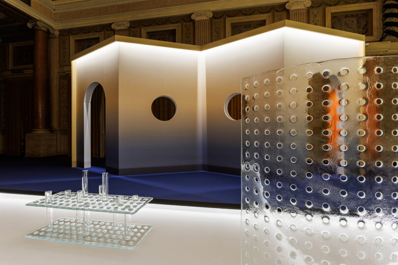 A modern interior with a geometric white structure featuring arched and circular openings, a perforated glass table, and a curved perforated glass partition.