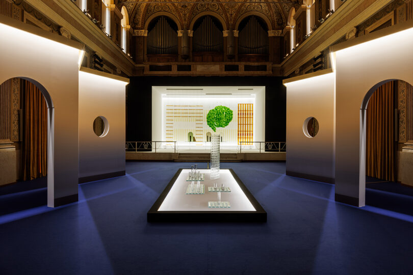A modern art installation with glass objects and a green topiary is displayed on a lit white platform in a grand hall with ornate arches and a decorative ceiling.