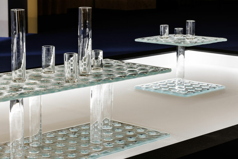 Two modern glass tables with bubble-like textures from the POETICA Wonderglass collection feature integrated cylindrical vases of varying heights, elegantly displayed on a lit surface.