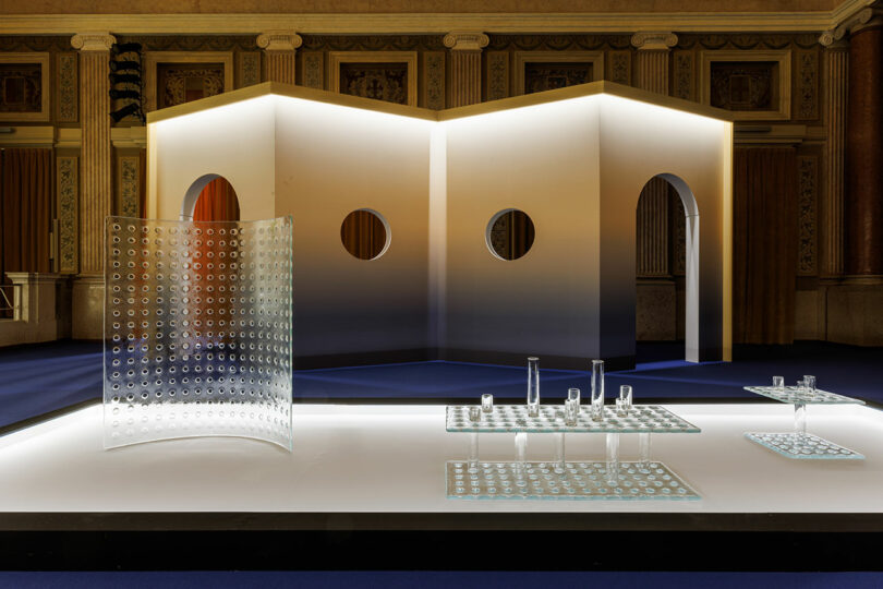 A modern exhibition space by POETICA Wonderglass features glass tables and cylinders on a raised platform, set against a curved wall with circular cutouts in the background.