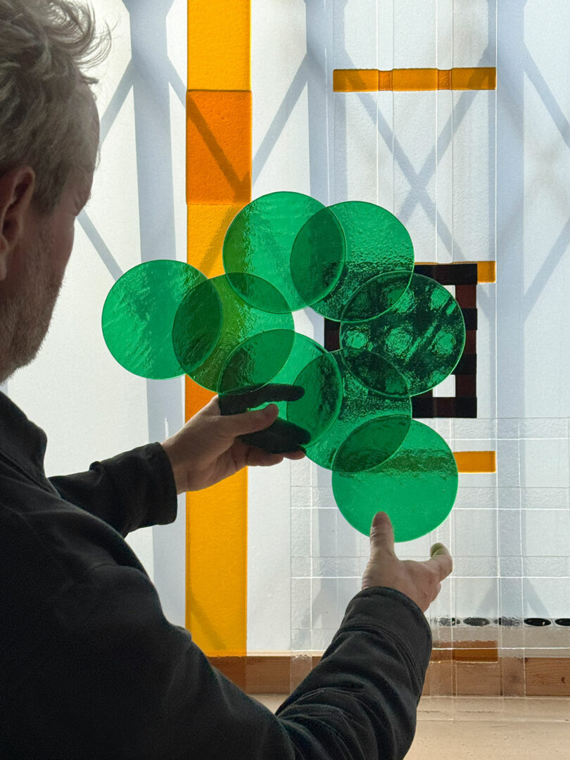 A person holds overlapping green glass discs from POETICA Wonderglass in front of a sunlit window with a geometric yellow, black, and white pattern.