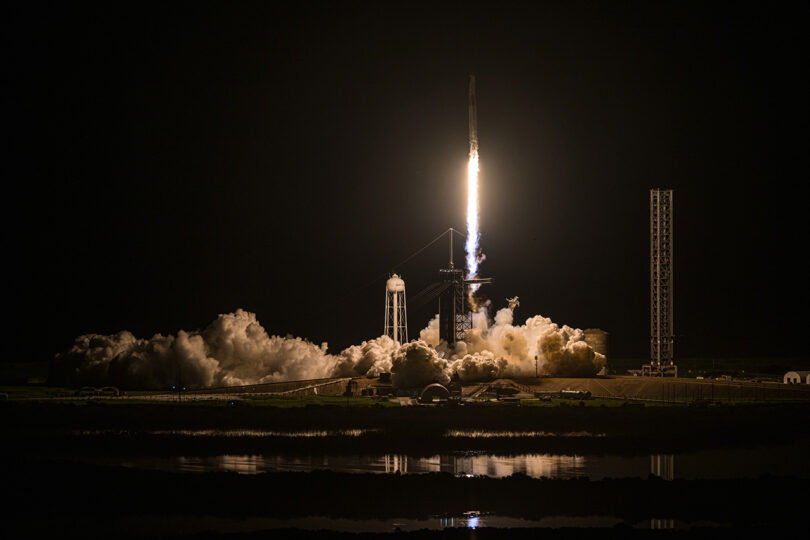 A rocket launches into the night sky, illuminating the surrounding area with bright flames and smoke, against a dark backdrop.