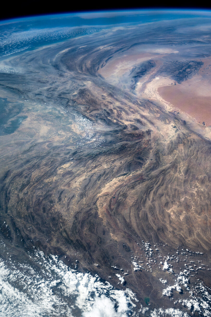 Aerial view of a vast desert landscape with rugged terrain and sparse cloud coverage, as seen from space.