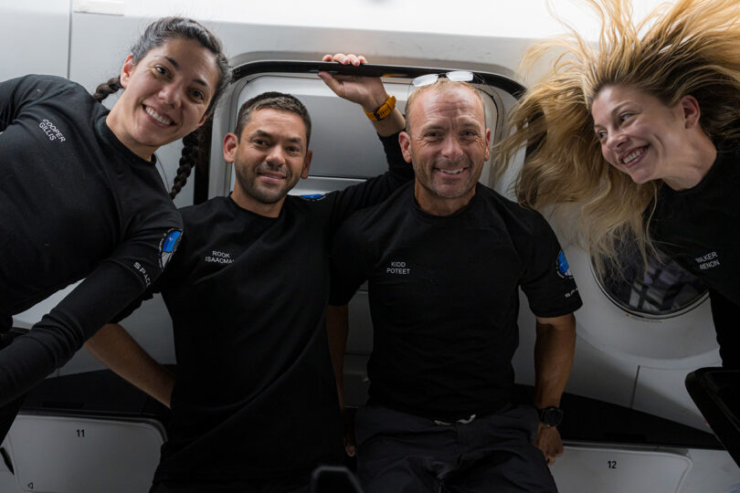Four astronauts in black uniforms float inside a spacecraft, smiling at the camera.