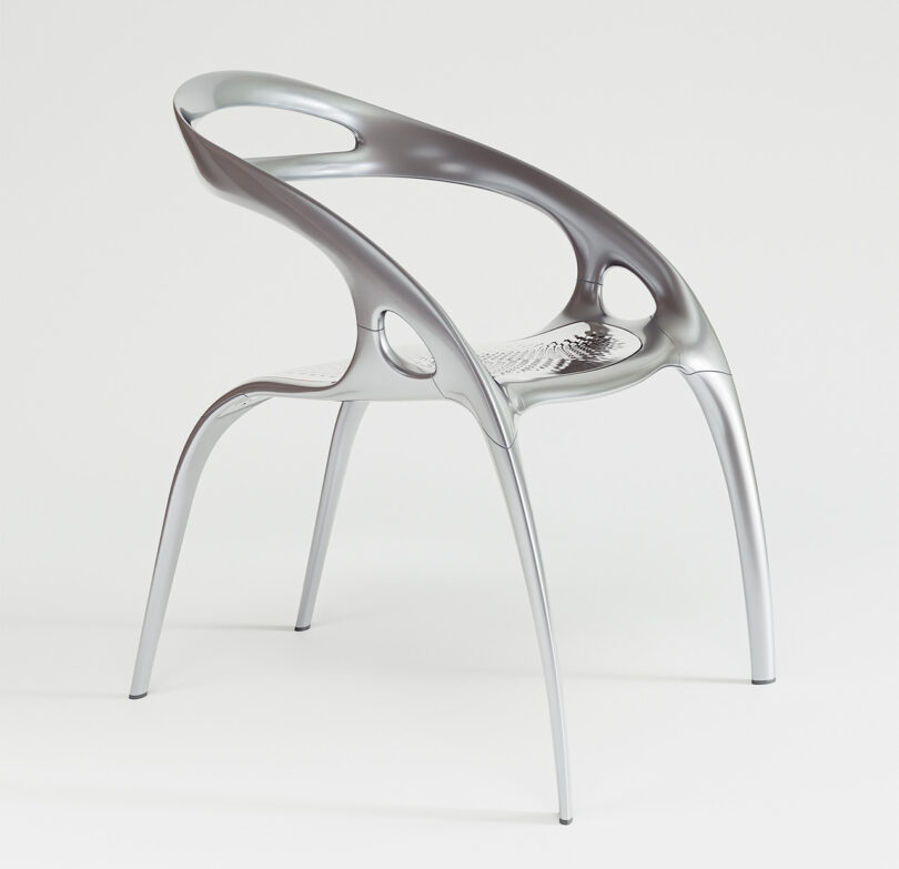 A sleek, modern metallic chair with a curved back and armrests, featuring circular cutouts. The chair has a minimalist design and stands on four slender legs.