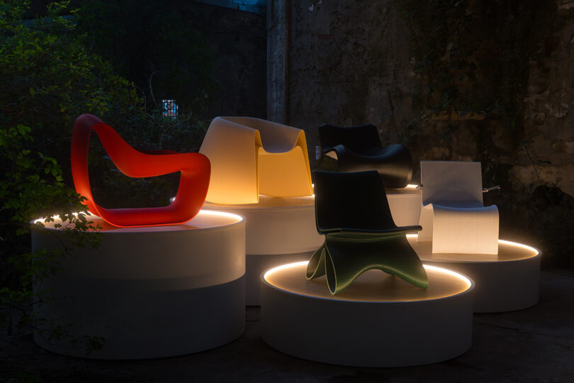 Five modern chairs in different colors and shapes are displayed on round, illuminated platforms in a dimly lit, industrial space.