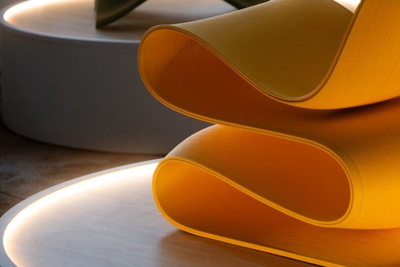 Close-up of a modern yellow lounge chair with curved, layered design, positioned on a round, lighted wooden platform.