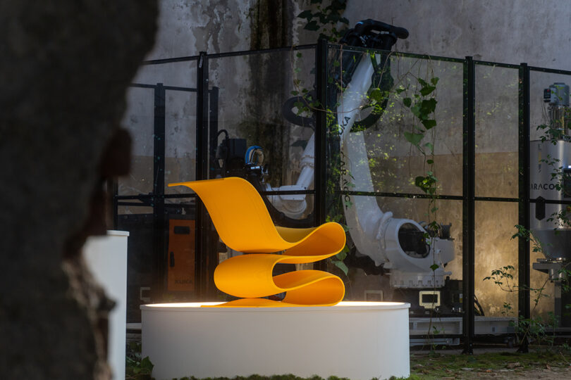 A bright yellow, modern, curved chair is displayed on a white platform in front of industrial robotic arms and a concrete wall.