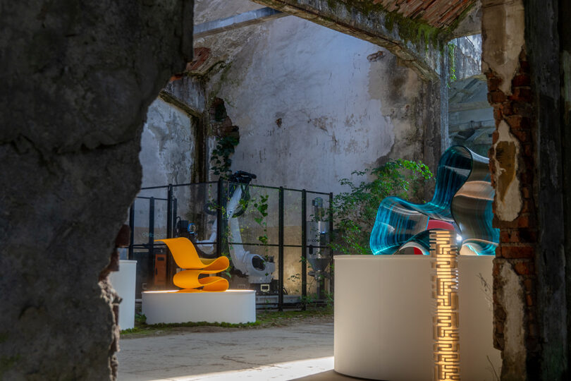 A display of modern furniture, including a yellow chair and blue transparent seats, is set inside a weathered, partially roofless building with exposed stone walls.