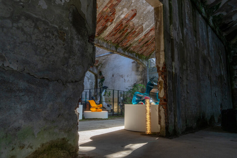 A view through a large hole in a weathered, partially roofless stone building reveals modern chairs and decor inside a sunlit, dilapidated interior.