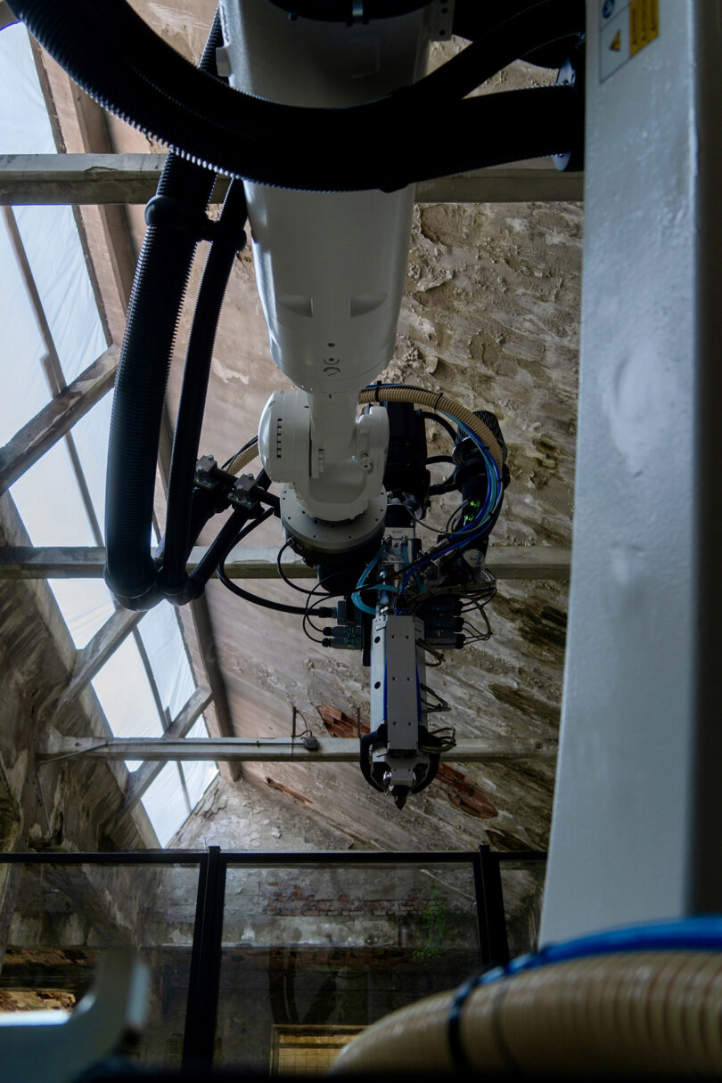 A large industrial robotic arm is suspended from the ceiling in a building with exposed stone walls and glass roof panels.