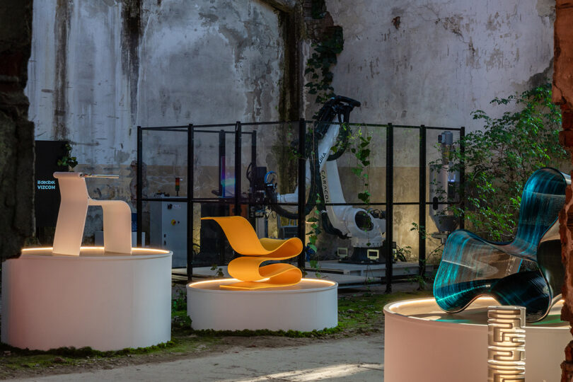 Three modern, sculptural chairs are displayed on round white platforms in an industrial space with exposed walls and a robotic arm in the background.