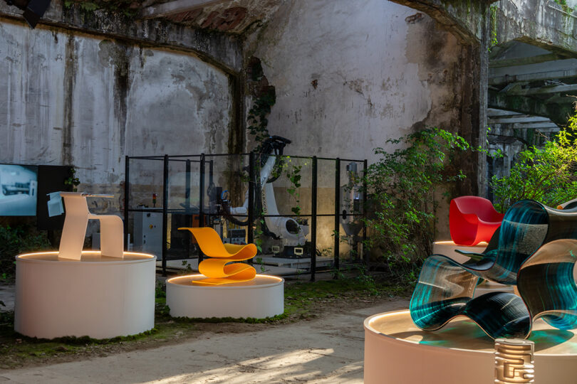 An industrial space with mossy walls displays modern chairs on white pedestals, with a robotic arm enclosed in glass and greenery growing around the area.