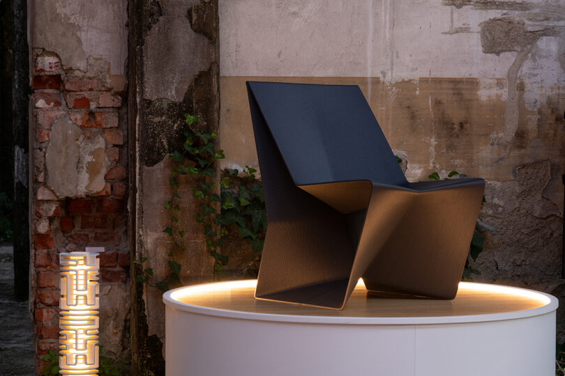 A modern black angular chair is displayed on a round, illuminated platform against a weathered wall with exposed bricks and climbing greenery.