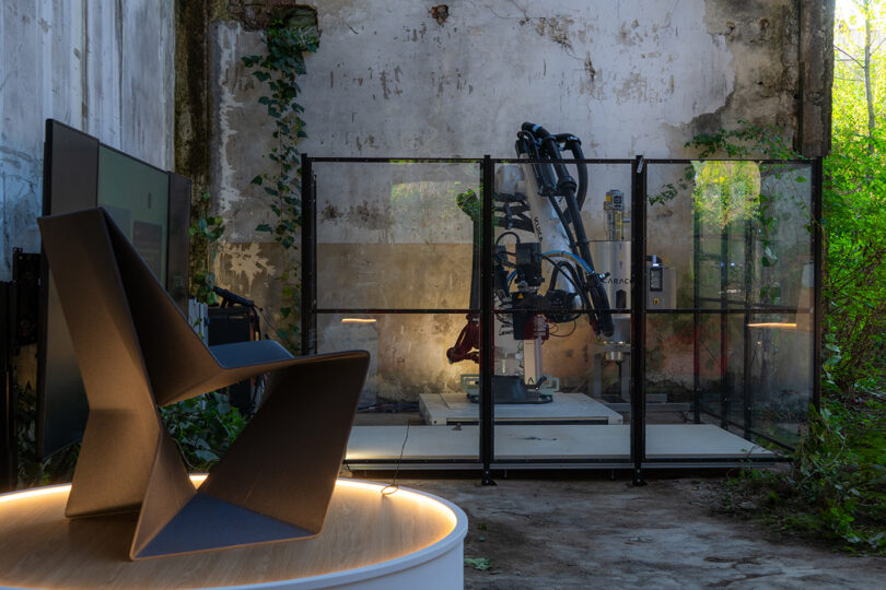 A modern black chair is displayed on a lit platform in the foreground; behind it, a robotic arm is enclosed in a transparent safety cage inside an old, weathered building with overgrown plants.
