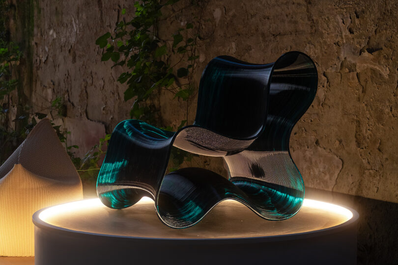 A sculptural, wavy chair with a glossy, dark green finish sits on a round, illuminated platform against a textured stone wall with some climbing plants.