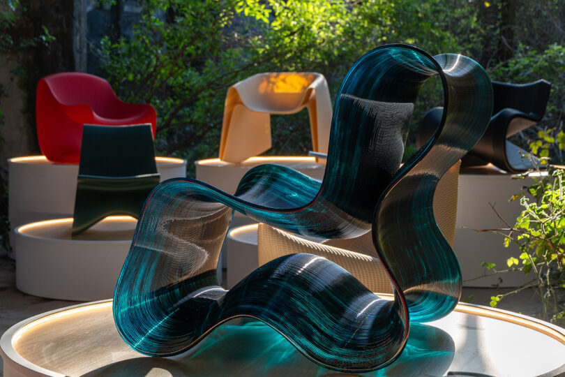 A wavy, translucent blue chair is displayed in the foreground, with four colorful modern chairs on lit circular platforms in the background amidst greenery.