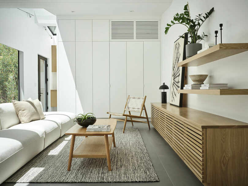 Minimalist living room with a white sofa, woven chair, wooden coffee table, wall shelves, and sideboard; neutral decor with natural light and green plant accents.