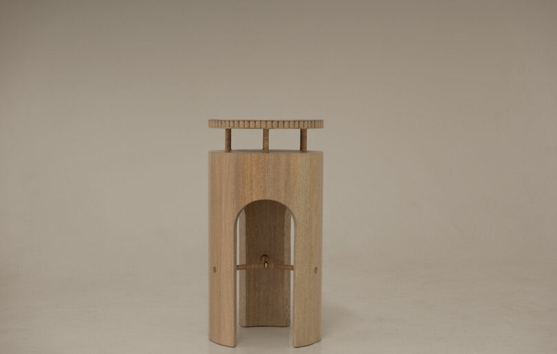 A wooden object with a curved base, central arched opening, and flat rectangular top supported by small vertical rods, set against a plain beige background—perfect for when you want to Take Five and admire simple design.