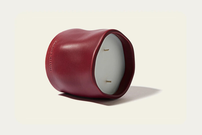 A red, irregularly shaped Take Five candle holder with a white candle and two wicks is shown lying on its side against a plain background.