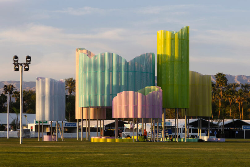 Large, colorful, translucent structures, in pastel shades, stand on a grassy field, with tents and palm trees - overcoming you and relaxed in the middle of this vibrant scene.