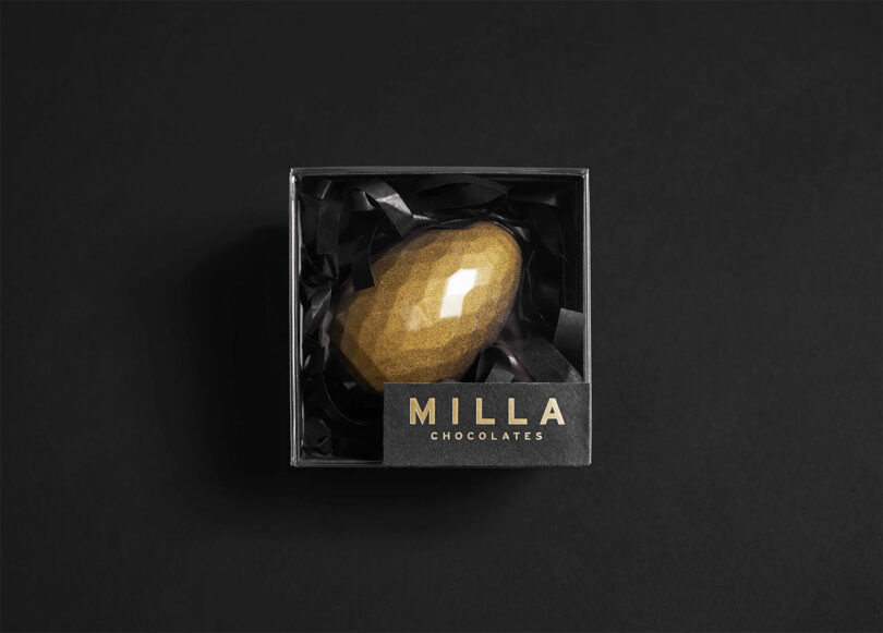 A gold, faceted, gem-shaped chocolate is nestled in black paper inside a black box labeled "Milla Chocolates" on a dark background.