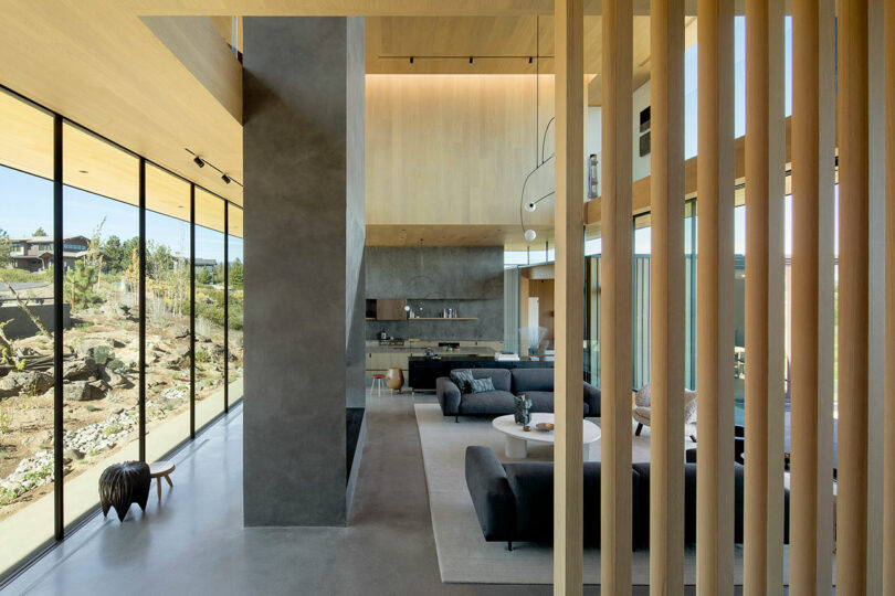 Modern open-plan living area with floor-to-ceiling windows, minimalist furniture, a gray accent wall, and a kitchen in the background. Natural light fills the space.