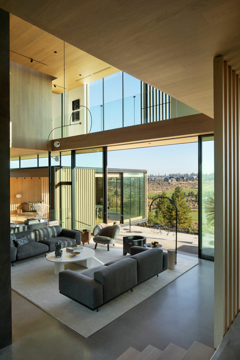 Modern living room with large windows, gray sofas, a white coffee table, and expansive views of an outdoor landscape under natural light.