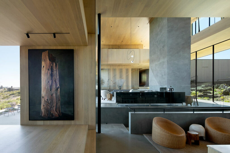 Modern open-plan living space with large windows, minimalist decor, wood and concrete surfaces, woven chairs, and a tall artwork featuring a tree trunk.