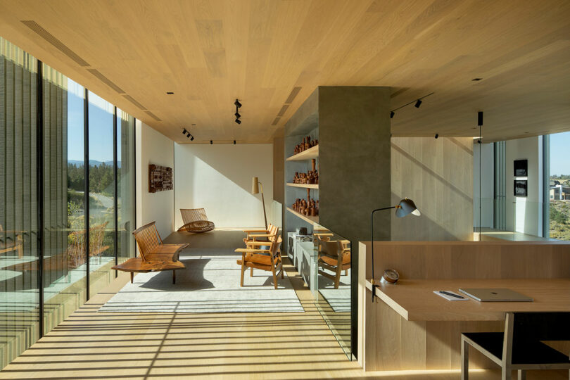 Modern office space with wooden ceiling and floors, large glass windows, minimalist furniture, built-in shelves, and a desk with a laptop and lamp. Natural light fills the room.